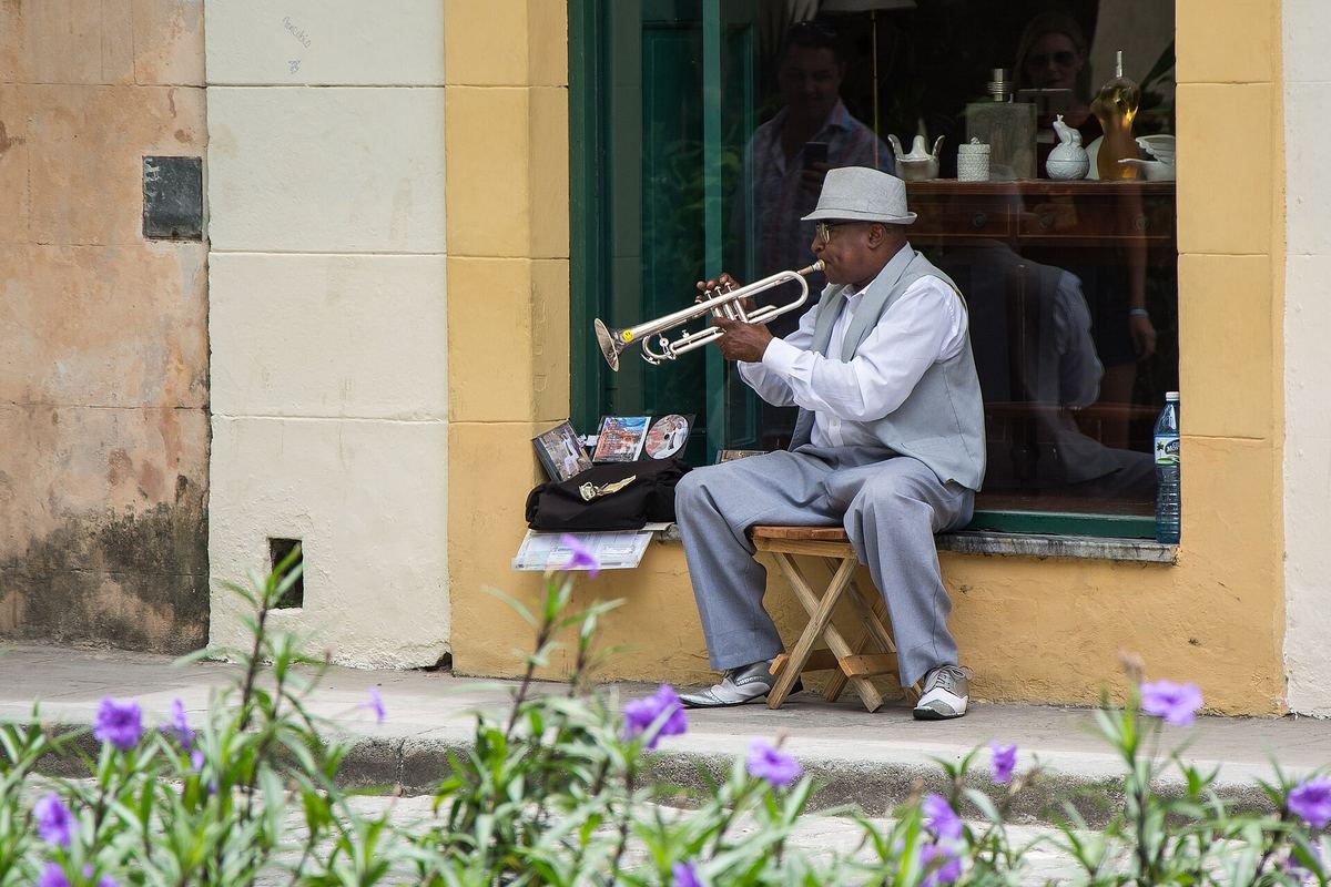 Ein älterer Mann mit weißem Hut sitzt vor einer Hauswand in Havanna und spielt Trompete